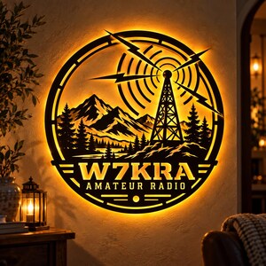 Puede incluir: Un letrero de pared circular de metal negro con un brillo naranja retroiluminado. El diseño muestra una torre de radio con rayos, montañas y pinos. El texto W7KRA AMATEUR RADIO se muestra claramente en la parte inferior del círculo.