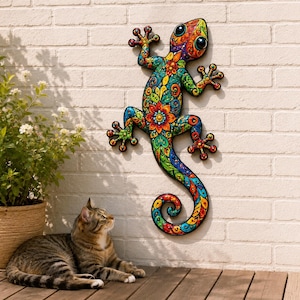Op de afbeelding: Een wandsculptuur van een gekko met een levendig, veelkleurig bloemen- en paisleypatroon. De hagedis is afgebeeld met grote ogen, gespreide tenen en een gekrulde staart, in een palet van heldere oranje, blauwe, groene en gele tinten.