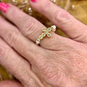 Peut inclure: Une bague ornée d'une croix dorée centrale sertie de petites pierres transparentes. L'anneau est composé d'une alternance de petites perles sphériques blanches et de minuscules entretoises métalliques dorées, formant un design circulaire délicat.