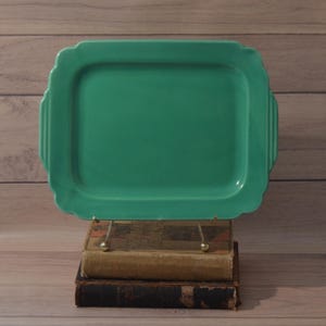 May include: A rectangular jade green serving tray with rounded corners and ribbed side handles. The tray features a smooth, glossy finish and a slightly raised edge, presenting a solid, uniform color throughout its entire surface area.