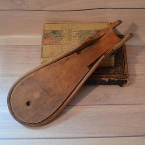 May include: A vintage wooden weaving shuttle with a smooth, light brown finish and a rounded end featuring a small circular hole. The shuttle has a long, tapered body with a central channel and is positioned diagonally across a weathered book.
