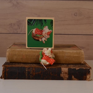May include: A small figurine of a white mouse wearing a red cape with white polka dots, nestled inside a brown walnut shell. The mouse is holding a smaller mouse figure. The figurine is displayed in front of its original square cardboard box.