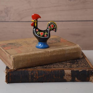 May include: A small black ceramic rooster figurine with a red comb and wattle, featuring hand-painted white dots, green leaves, and red heart motifs on its body. The base is blue with the word Portugal written in white script lettering.