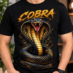 Könnte beinhalten: Ein schwarzes Kurzarm-T-Shirt mit einem großen Grafikdruck einer zusammengerollten Kobra mit gespreizter Haube, orangefarbenen Augen und offenem Maul mit Fangzähnen. Das Wort COBRA ist in einer goldgelben Schrift über der Schlange abgebildet.
