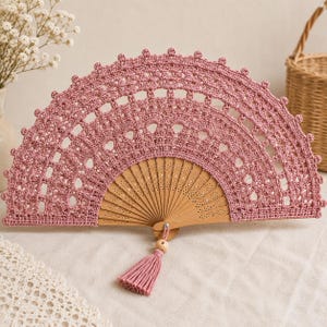 May include: A handheld folding fan featuring a light pink crocheted lace cover with an intricate openwork pattern. The fan has a natural light brown wooden frame with perforated details and a matching pink tassel attached by a small wooden bead.