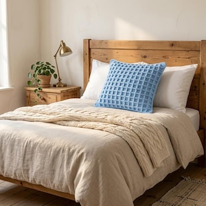 May include: A square light blue waffle-knit throw pillow centered on a bed. The pillow features a raised grid texture and is positioned in front of two plain white pillows. The bed is covered with a beige linen duvet and a cream cable-knit blanket.