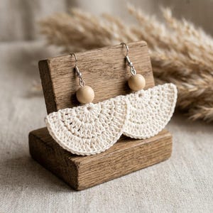 Puede incluir: Un par de pendientes de ganchillo en color blanco roto con forma de abanico semicircular suspendidos de una cuenta de madera clara. Los pendientes están sujetos a ganchos de metal plateado y muestran un patrón de punto de hilo texturizado.