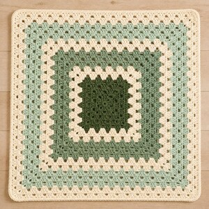 May include: A square crochet blanket featuring a classic granny square pattern. The design consists of concentric layers in alternating shades of cream, light sage green, and a deep forest green, creating a geometric, multi-coloured textile piece.