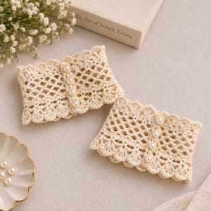 May include: A pair of cream-colored crochet wrist cuffs featuring an openwork diamond pattern and a vertical row of four white faux pearls on each piece. The edges of the cuffs have a scalloped design, and the material is a soft yarn.