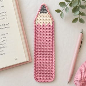 Op de afbeelding: Een gehaakte boekenlegger in de vorm van een potlood met een roze lichaam, een crèmekleurig gedeelte dat het hout voorstelt en een donkergrijze punt. De boekenlegger heeft een gestructureerd, gehaakt patroon en ligt verticaal op een ondergrond.