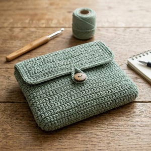 May include: A sage green crocheted pouch featuring a rectangular flap secured by a single round wooden button. The pouch is crafted with a dense, uniform stitch pattern and is positioned alongside a wooden crochet hook and a spool of matching yarn.