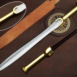 May include: A double-edged sword with a polished steel blade and a gold-toned metal handle featuring a distinctive flared pommel and guard. The set includes a long, tan leather scabbard and a dark brown shield-shaped plaque with a gold Greek key pattern.