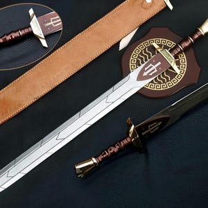 Könnte beinhalten: Ein zweischneidiges Schwert mit silberner Klinge, geometrischen Gravuren, dunkelbraunem Griff sowie goldfarbenem Knauf und Parierstange. Dazu gehören eine beigefarbene Lederscheide und eine braune, schildförmige Plakette mit goldenem Mäander-Rand.