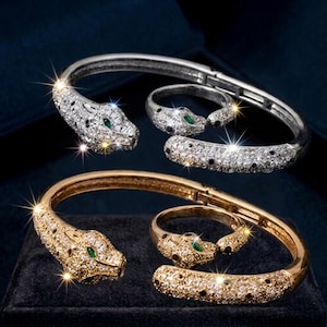 Könnte beinhalten: Zwei Schmucksets, bestehend aus jeweils einem aufklappbaren Armreif und einem passenden Ring. Ein Set ist silberfarben, das andere goldfarben. Beide zeigen ein Pantherkopf-Design, besetzt mit klaren Kristallen, schwarzen Punkten und grünen Augen.