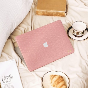 Puede incluir: Una funda para portátil de color rosa con un patrón texturizado de piel de cocodrilo y un recorte para el logotipo de Apple en color plata. La funda presenta iniciales monogramadas en color dorado en la esquina inferior derecha.
