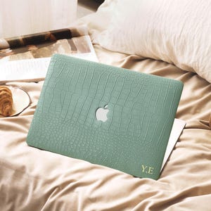 May include: A sage green laptop case with a textured crocodile skin pattern and a cutout for the Apple logo. The case features the gold-coloured initials Y.E. printed in the bottom right corner, providing a protective cover for the laptop device.