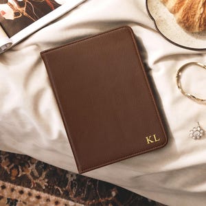 May include: A rectangular brown leather notebook with a textured surface and rounded corners. The bottom right corner features the gold embossed initials KL. The notebook is closed and has a clean, minimalist design with visible stitching along the edges.