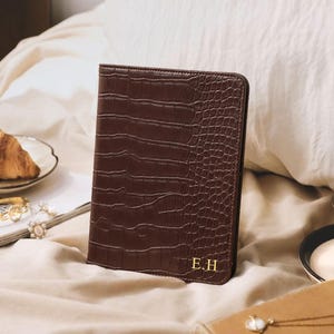May include: A dark brown rectangular passport holder with a textured crocodile skin pattern. The front cover features the gold embossed initials E.H. in the bottom right corner. The item is a vertical, book-style case with stitched edges.