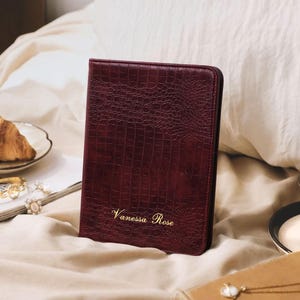 May include: A dark burgundy, rectangular notebook cover with a textured crocodile skin pattern. The front features the name Vanessa Rose embossed in gold lettering. The cover is upright, showcasing a structured spine and a smooth, finished edge.