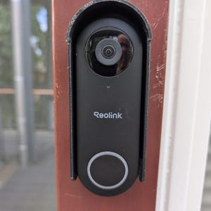 Op de afbeelding: Een zwarte Reolink videodeurbel gemonteerd op een roodbruin deurkozijn. Het apparaat is voorzien van een ronde cameralens aan de bovenkant, de merknaam Reolink in het wit in het midden en een grote ronde knop met een lichtring aan de onderkant.
