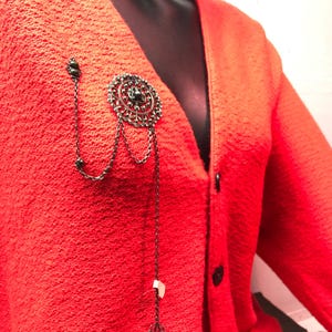 Peut inclure: Un cardigan orange vif à texture granuleuse orné d'une broche en métal foncé au motif filigrane circulaire. La broche est agrémentée de plusieurs chaînes délicates qui tombent vers le bas, terminées par de petites perles sombres et une étiquette.