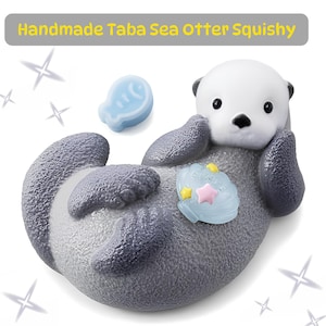 May include: A handmade Taba sea otter squishy toy with a white face, dark gray body, and textured surface. The otter holds a light blue seashell with small yellow and pink stars, accompanied by a separate light blue fish-shaped piece.