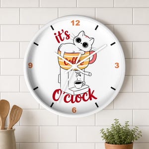 May include: A round white wall clock featuring a cartoon illustration of a white cat resting in a striped coffee mug atop a coffee machine. The text reads it's coffee O'clock in red. The clock has black hour markers and orange numbers 3, 6, 9, 12.