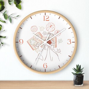 May include: A round wall clock with a light wood frame and a white face. The design features orange line art of pumpkins, maple leaves, an open book, a camera, and hands holding a mug. The numbers 3, 6, 9, and 12 are printed in orange.
