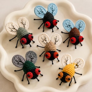 May include: Six small, handcrafted crochet insects arranged on a scalloped white tray. Each figure features a round body in various colors including black, green, brown, and yellow, paired with light blue or cream wings and prominent red or orange eyes.