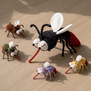 Puede incluir: Una colección de cinco juguetes de mosquito tejidos a ganchillo en varios colores, incluyendo negro, marrón, verde, morado y amarillo. Cada juguete presenta grandes ojos blancos con pupilas negras, patas finas, alas blancas y una trompa roja.
