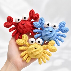 以下が含まれることがあります： 鮮やかな赤、水色、黄色の3つの小さなかぎ針編みのカニのおもちゃ。各カニは丸い体、2つの大きなハサミ、複数の細い足、黒い瞳を持つ2つの白い目、そして黒い糸で刺繍された口が特徴です。