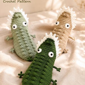 Puede incluir: Tres juguetes de cocodrilo tejidos a mano en verde salvia, verde oscuro y beige. Cada uno presenta una cresta blanca con puntas en la espalda, extremidades pequeñas y ojos negros y blancos. El texto PDF Crochet Pattern es visible arriba.