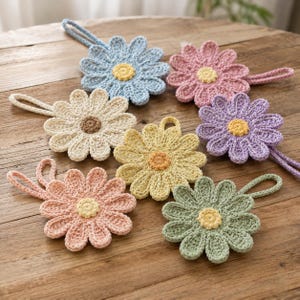 Può includere: Una collezione di sette ornamenti a forma di fiore all'uncinetto in vari colori, tra cui azzurro, rosa, crema, giallo, lavanda, pesca e verde salvia. Ogni fiore presenta un dettaglio circolare centrale e un occhiello per appenderlo.