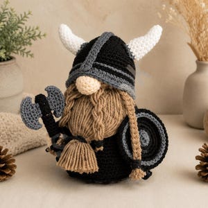 May include: A crocheted gnome figure wearing a black and gray Viking helmet with white horns. The gnome has a tan yarn nose, a long braided tan beard, and is holding a gray double-bladed axe in one hand and a round black and gray shield in the other.