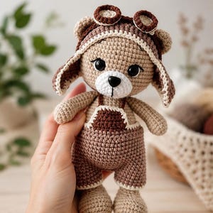 May include: A crocheted teddy bear toy featuring light brown yarn, a white muzzle, and a black nose. The bear wears dark brown overalls and a matching aviator-style cap with two circular goggles on top, all crafted with detailed stitching.