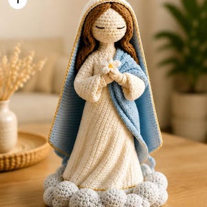 Puede incluir: Una muñeca de ganchillo que representa una figura con cabello largo castaño, vestida con una túnica color crema y una capa con capucha azul claro con borde dorado. La figura se apoya sobre una base de nubes grises y sostiene una flor blanca.