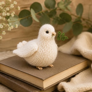 May include: A small, cream-colored knitted bird figurine with a soft, fuzzy texture. The bird has a round body, a small beak, and dark eyes. It holds a tiny, green crocheted four-leaf clover in its beak, adding a touch of color to the design.