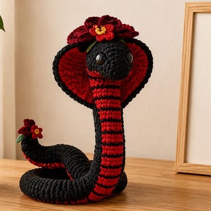 May include: A crocheted cobra toy featuring a black and red striped body, a flared hood, and dark eyes. The snake is adorned with small red and yellow crocheted flowers on its head and tail. Beside it sits a framed sign with the text BE KIND BE BRAVE BE YOU.