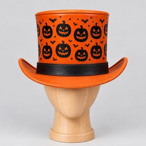 Puede incluir: Un sombrero de copa naranja con un patrón negro repetitivo de calabazas de Halloween, pequeños murciélagos y estrellas. El sombrero tiene un ala ancha y curva, y una banda negra sólida que rodea la base de la copa con un acabado liso.