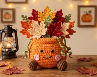 May include: A crocheted orange pumpkin-shaped container with a smiling face, pink cheeks, and two brown feet. The pumpkin is filled with a vibrant arrangement of crocheted autumn leaves in shades of red, yellow, orange, cream, and green, featuring curly green vines.