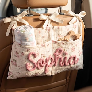 May include: A cream-colored fabric car seat organizer featuring a delicate pink and green floral pattern. The organizer has two pockets, cream ribbon ties for attachment, and the name Sophia embroidered in raised pink yarn across the front.