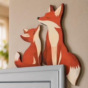 May include: Two flat, cutout wooden fox figures in a reddish-orange hue with cream-coloured chest and tail accents. The larger fox stands upright while the smaller fox sits beside it, both featuring closed eyes and simple, clean black line details.