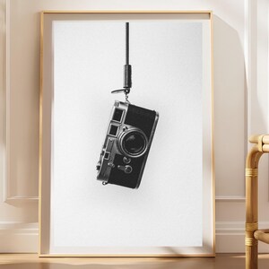 Peut inclure: Une impression d'art mural encadrée en noir et blanc représentant un appareil photo télémétrique de style vintage suspendu verticalement par une fine sangle noire sur un fond blanc uni. Le boîtier est noir avec des détails argentés.