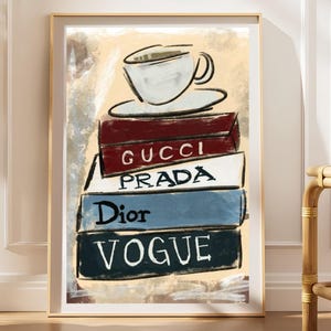 May include: A framed wall art print featuring a stack of four books topped with a coffee cup. The books are labeled GUCCI in red, PRADA in white, Dior in blue, and VOGUE in dark gray. The illustration uses visible brushstrokes on a beige background.
