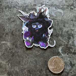 May include: A sticker featuring a black Furby-like creature with large blue eyes, adorned with feathers and purple accents. The sticker is placed on a textured, dark gray surface next to a U.S. quarter.