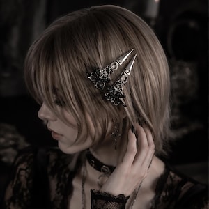 May include: Two silver-toned hair clips featuring intricate filigree metalwork and small black decorative accents. The clips have a pointed, dagger-like shape and are secured in light brown hair, positioned side-by-side above the ear.
