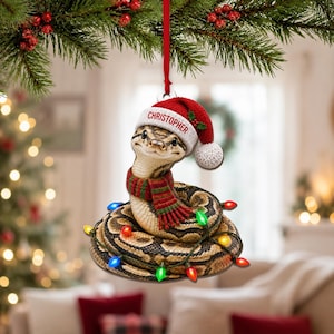 May include: A hanging Christmas ornament featuring a coiled brown and tan patterned snake wearing a red and white Santa hat with the name CHRISTOPHER printed on the brim, a red and green striped scarf, and wrapped in colorful string lights.