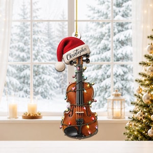 May include: A violin-shaped hanging ornament featuring a red Santa hat with the name Christopher printed on the white trim. The instrument is wrapped in a string of multicolored Christmas lights, including red, green, and blue bulbs.