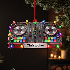 Könnte beinhalten: Ein rotes DJ-Controller-Ornament mit zwei kreisförmigen Jogwheels, bunten quadratischen Pads und einem zentralen Mixer-Bereich. Der Name Christopher ist auf einer silbernen Platte unten aufgedruckt. Eine bunte Lichterkette ist darum gewickelt.