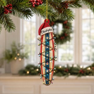 May include: A surfboard-shaped Christmas ornament featuring a red, white, and blue striped design with white stars. The ornament is wrapped in a string of colourful Christmas lights and topped with a red Santa hat bearing the name Christopher.